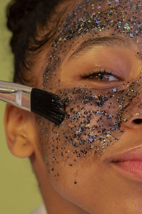 Free Crop unrecognizable young African American female with brush applying facial mask with glitters during beauty procedure and looking at camera on green background Stock Photo