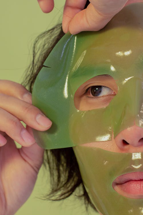 Crop unrecognizable Asian male with dark hair taking off facial mask during daily skincare procedure on green background in light studio