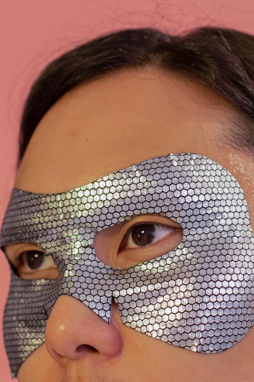 Crop unrecognizable Asian male with dark hair and gray eye mask standing on pink background during skincare procedure in studio