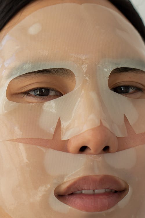 Unrecognizable Asian male with dark hair and moisturizing mask on face looking at camera during cosmetic procedure in light room