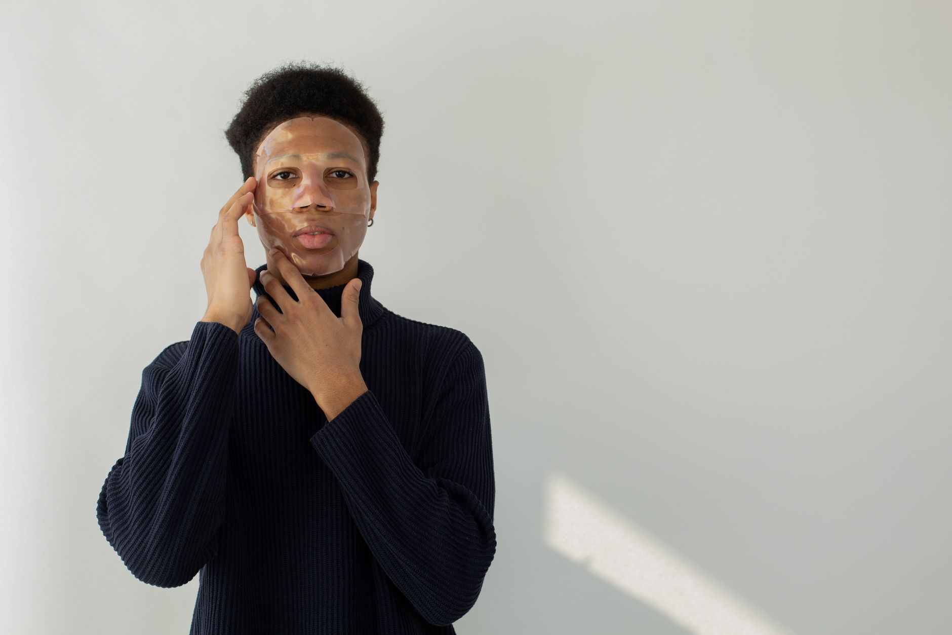Calm young African American male with moisturizing mask on face looking at camera on white background during skincare routine in studio