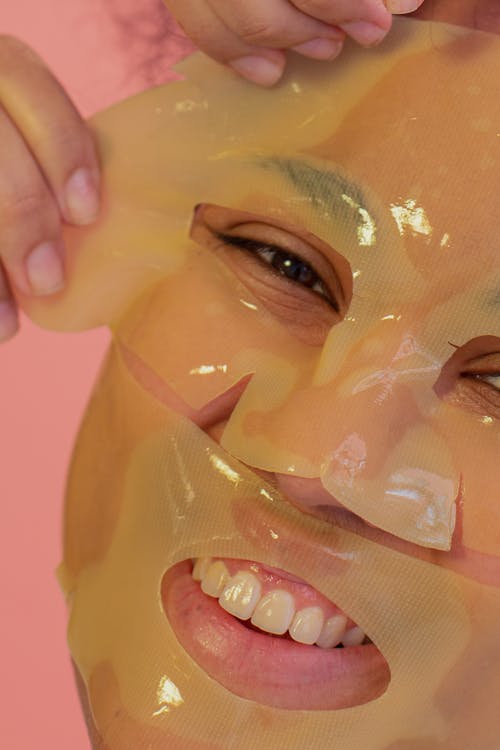Crop unrecognizable young female taking off yellow moisturizing facial mask and looking at camera during beauty procedure on pink background in studio