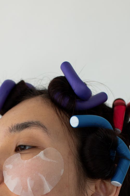 Crop anonymous Asian female with moisturizing eye patch and curlers in hair standing on white background during beauty procedure in studio