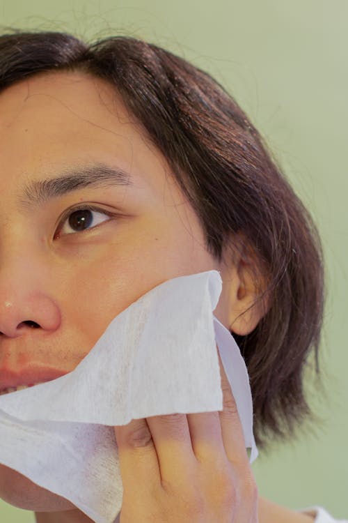 Crop young Asian male wiping face with wet wipe after cosmetic procedure while standing on green background in light studio