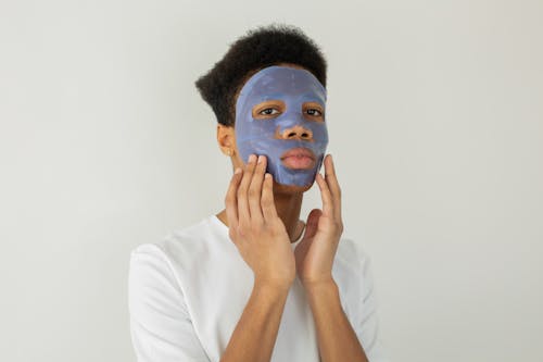 Serious young African American male with moisturizing facial mask applied on face looking away while standing on white background in studio