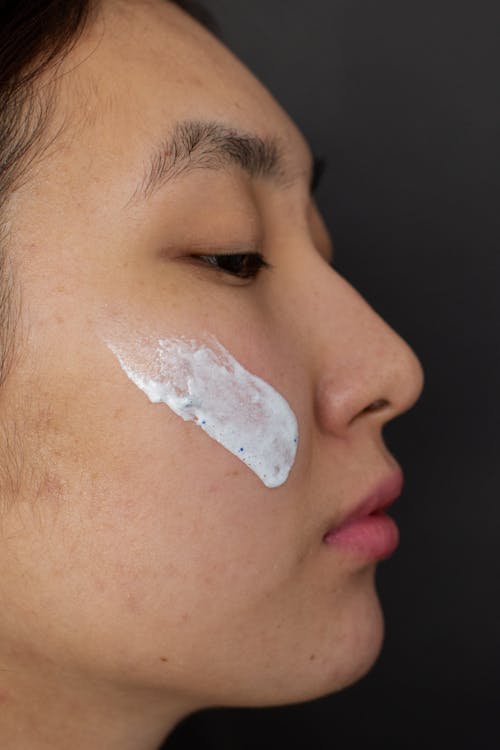 Side view of calm Asian male with smear of white clay mask standing on black background during skincare routine in studio