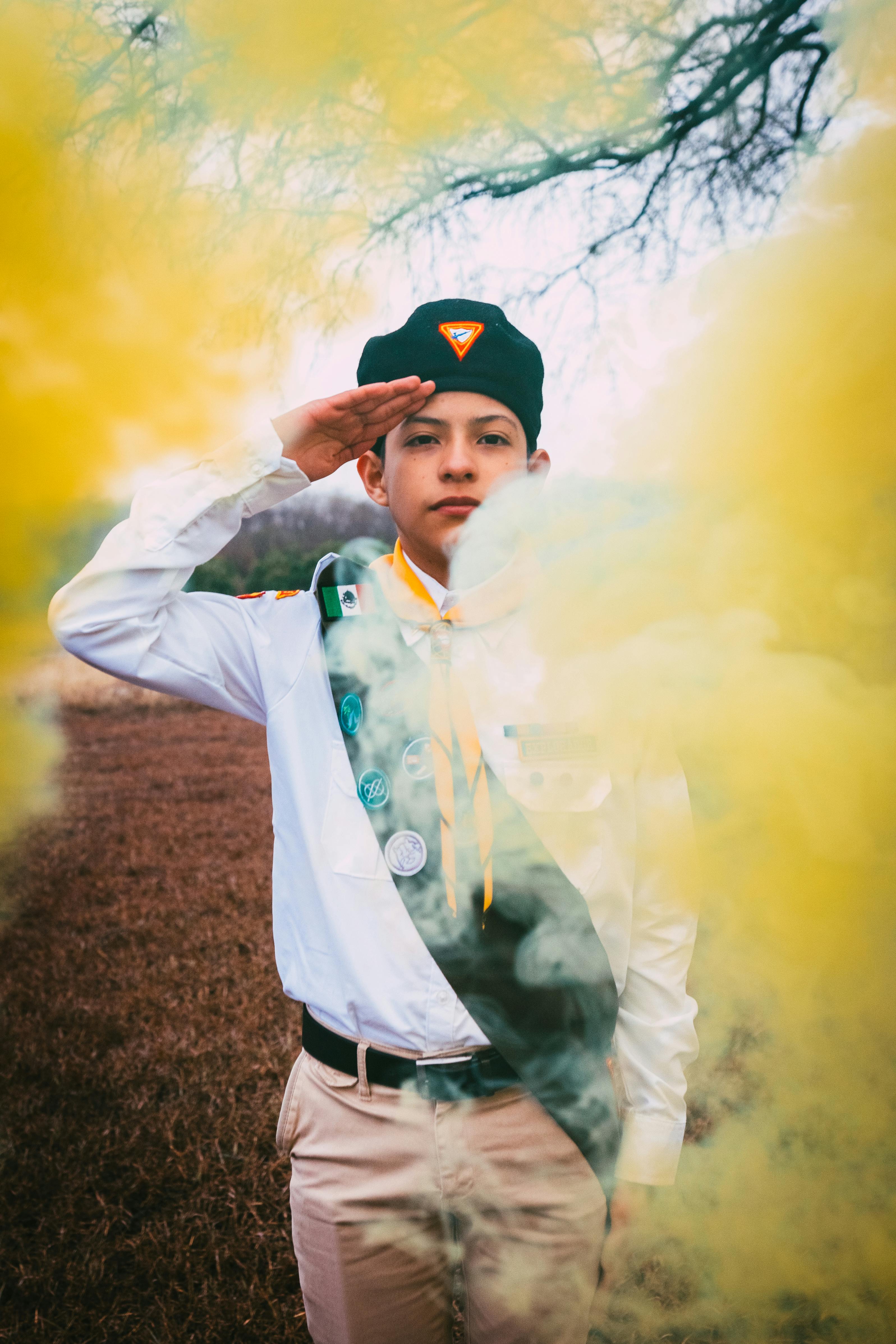 A young scout in uniform salutes surrounded by vibrant yellow smoke outdoors.