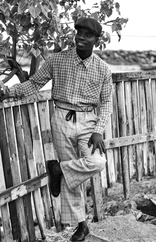 Grayscale Photo of a Man Posing Beside a Wooden Fence