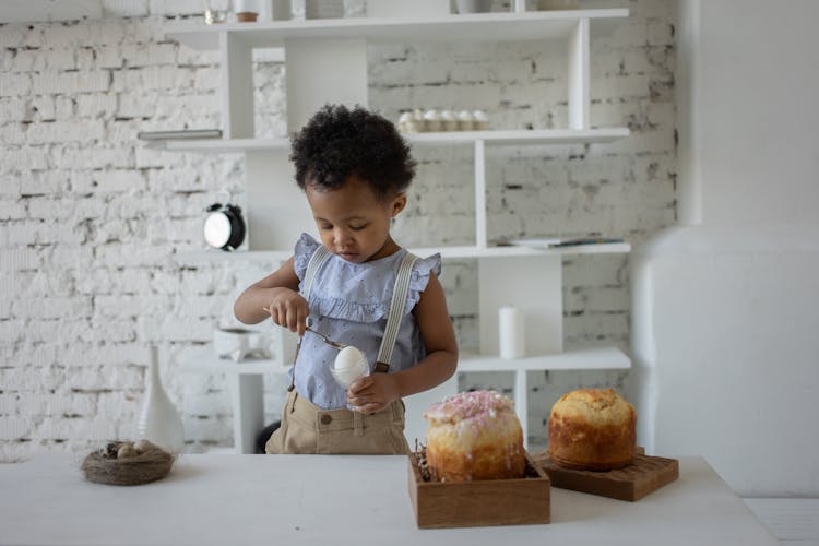 Little Girl Standing In A Kitchen And Tapping An Egg With A Spoon 