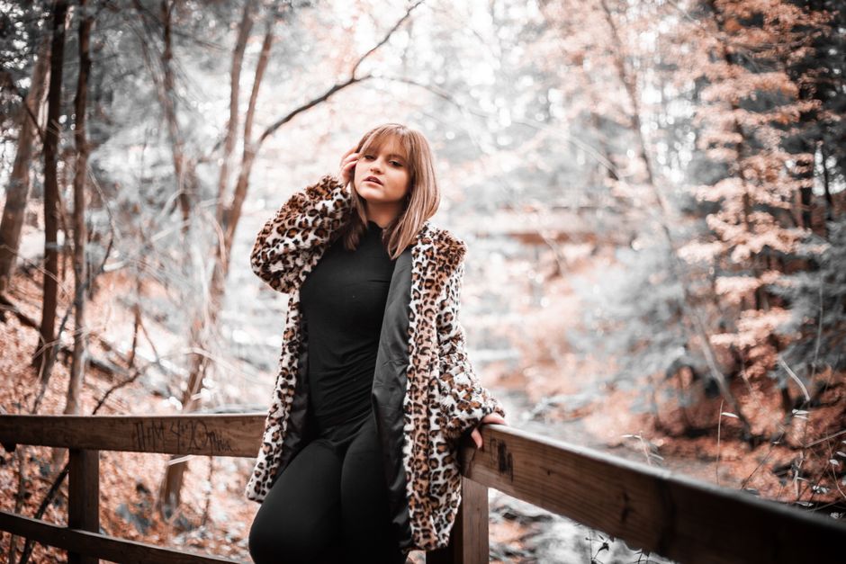 Stylish woman in leopard print coat posing in a fall forest setting.