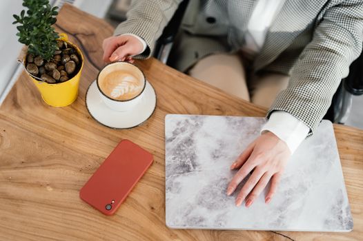 From above of unrecognizable female freelancer in wheelchair sitting at table with hot coffee and closed netbook during remote work in cafe