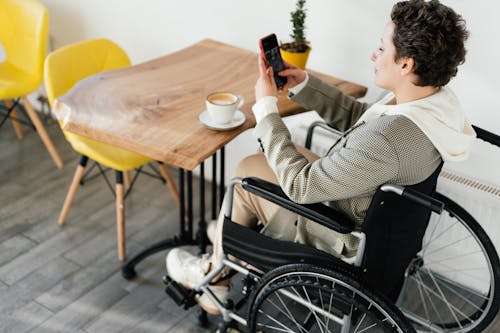 Free Side view full body of female in wheelchair taking picture of hot coffee while sitting at wooden table in cafeteria Stock Photo