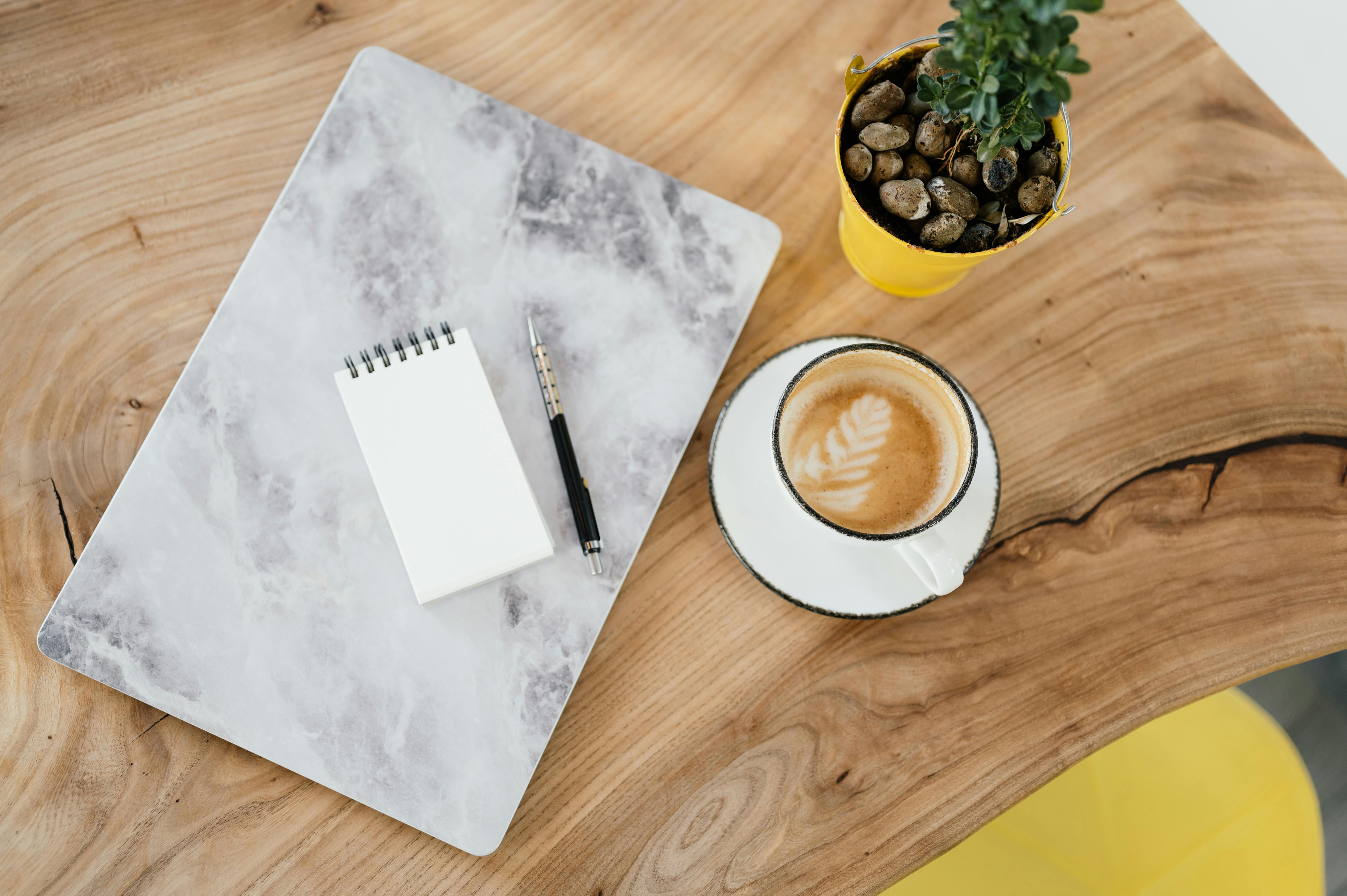Free Top view cup of hot latte on wooden table near modern laptop and notebook with pen placed near potted plant in cafe Stock Photo