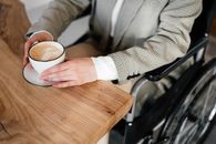 From above of unrecognizable female sitting in wheelchair at wooden table with cup of latte while spending time in cafe