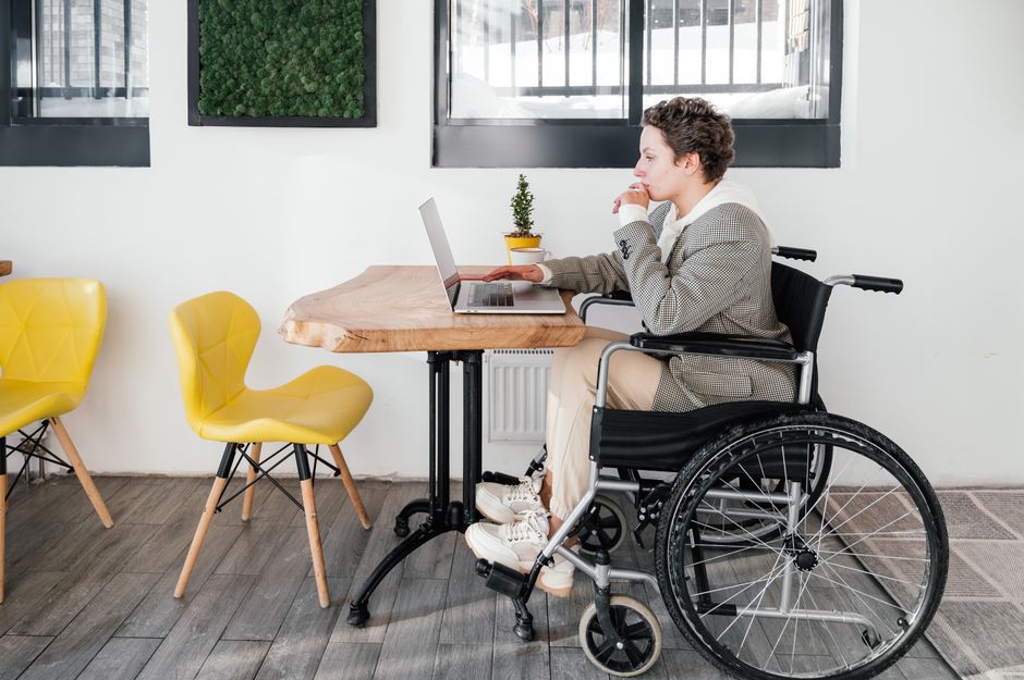 Full body side view of focused disabled female freelancer browsing internet on netbook while sitting at table during online work in cafeteria