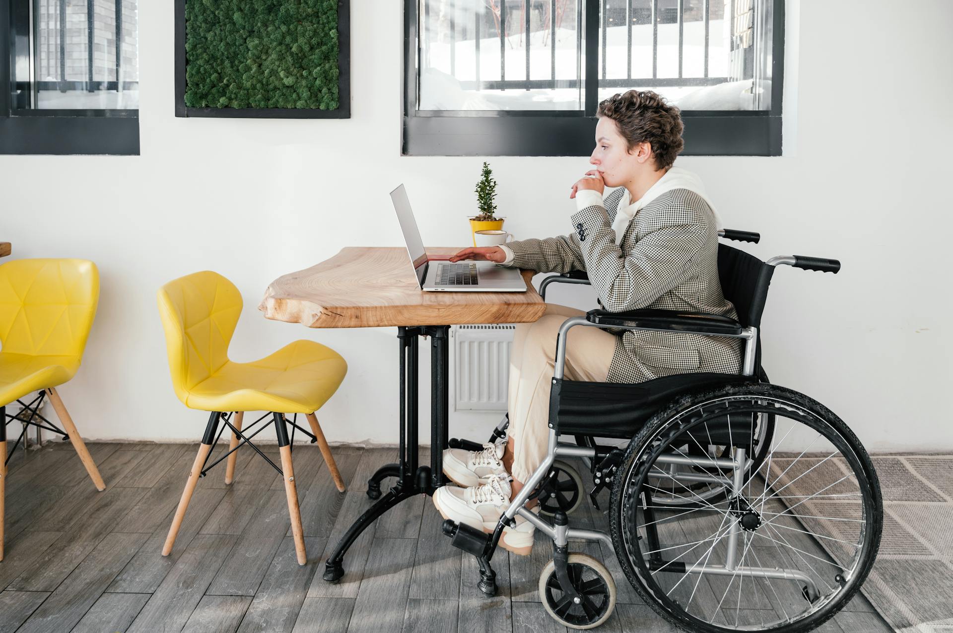 Full body side view of focused disabled female freelancer browsing internet on netbook while sitting at table during online work in cafeteria