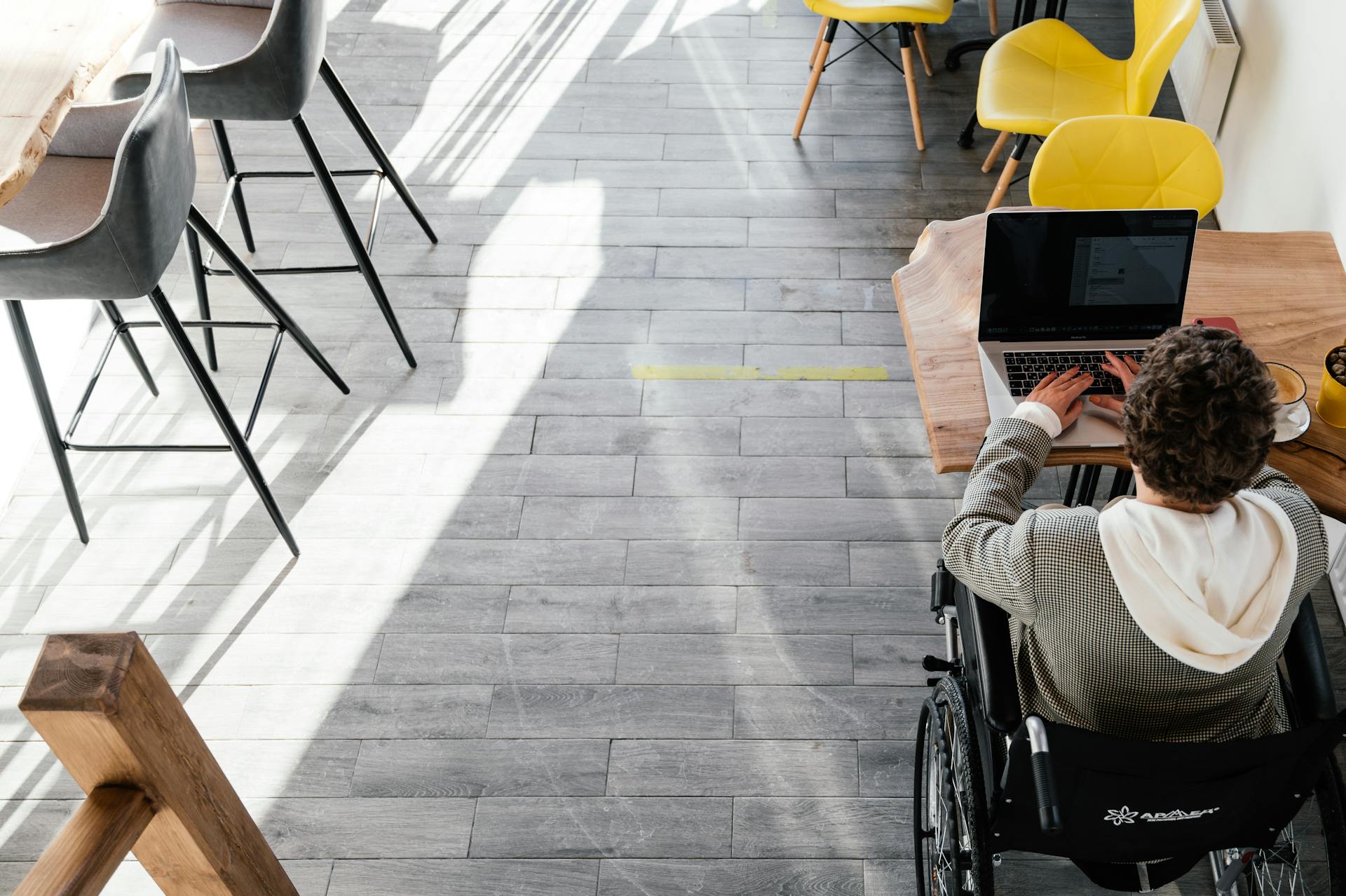 From above of anonymous disabled female freelancer typing on netbook sitting at table while working remotely in modern spacious cafeteria