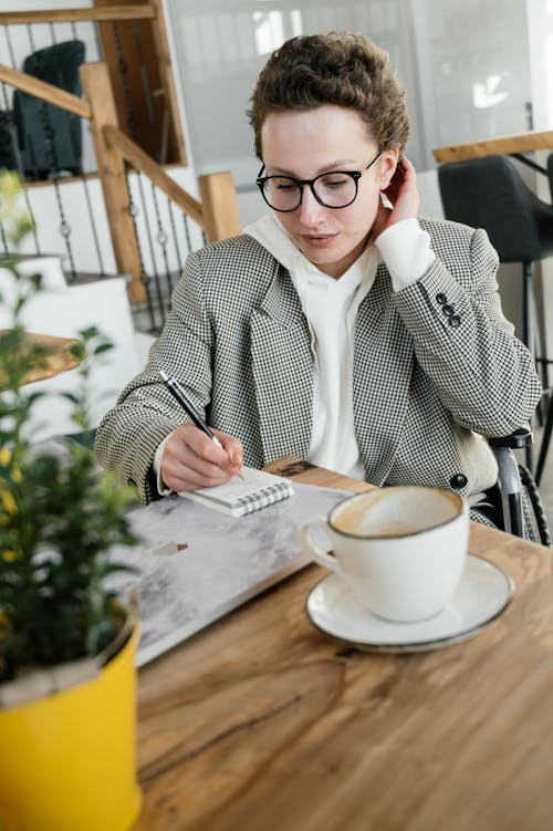 High angle of concentrated female in wheelchair writing in notebook while sitting at table with laptop and coffee