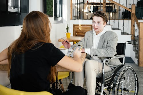 Concentrated female using wheelchair talking to coworker during coffee break in modern cafeteria