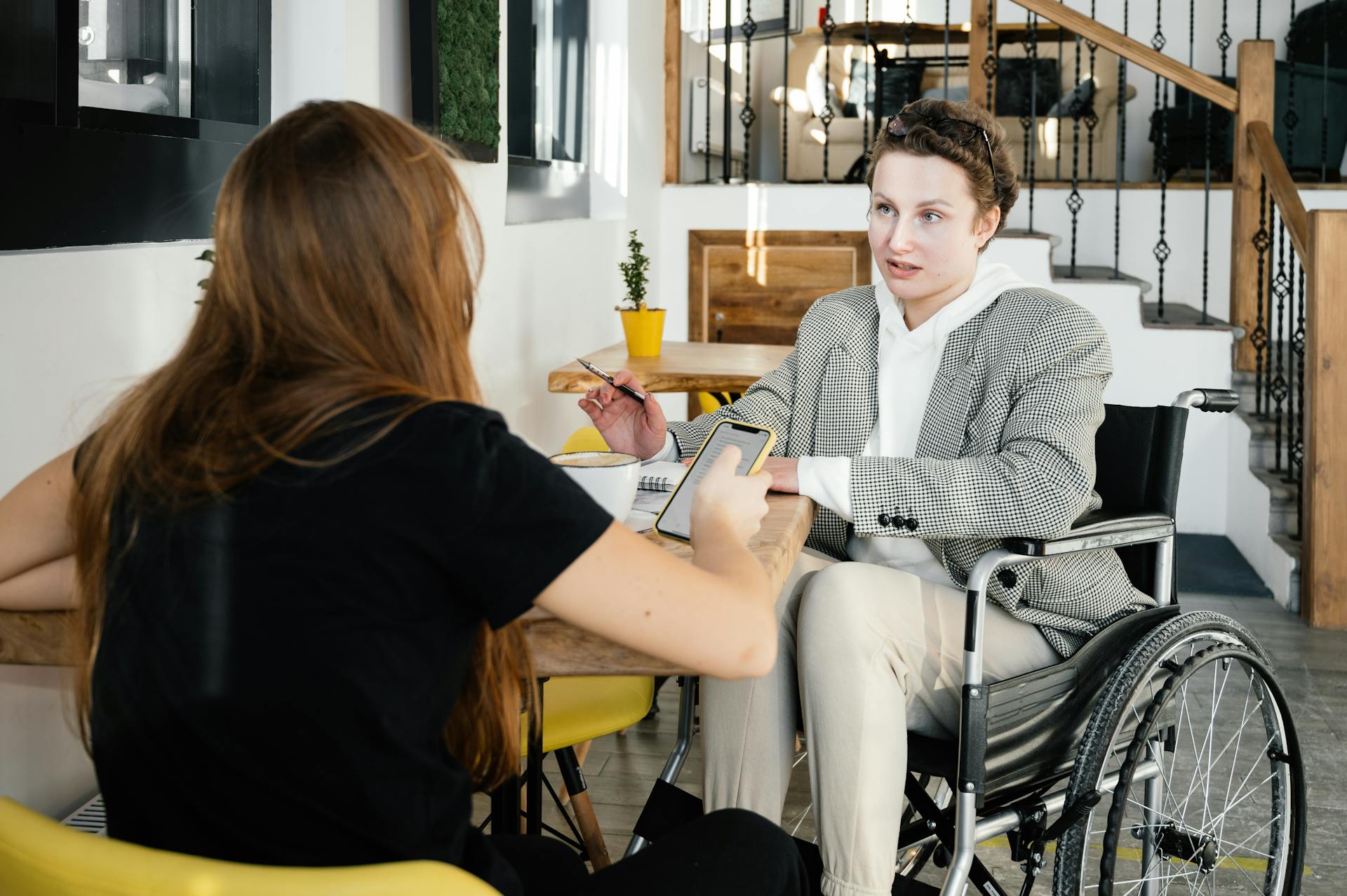 A person in a wheelchair having a conversation with another individual across a wooden table in a cozy, modern café environment.