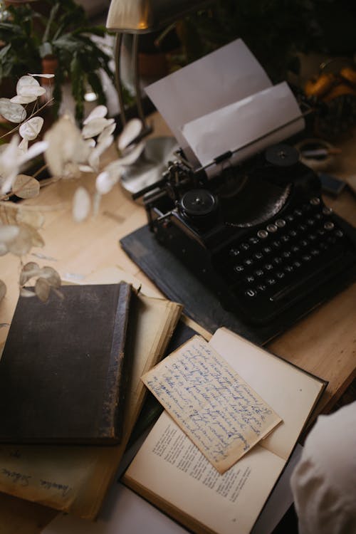 A Black Typewriter with a Bond Paper Beside a Book on a Wooden Table