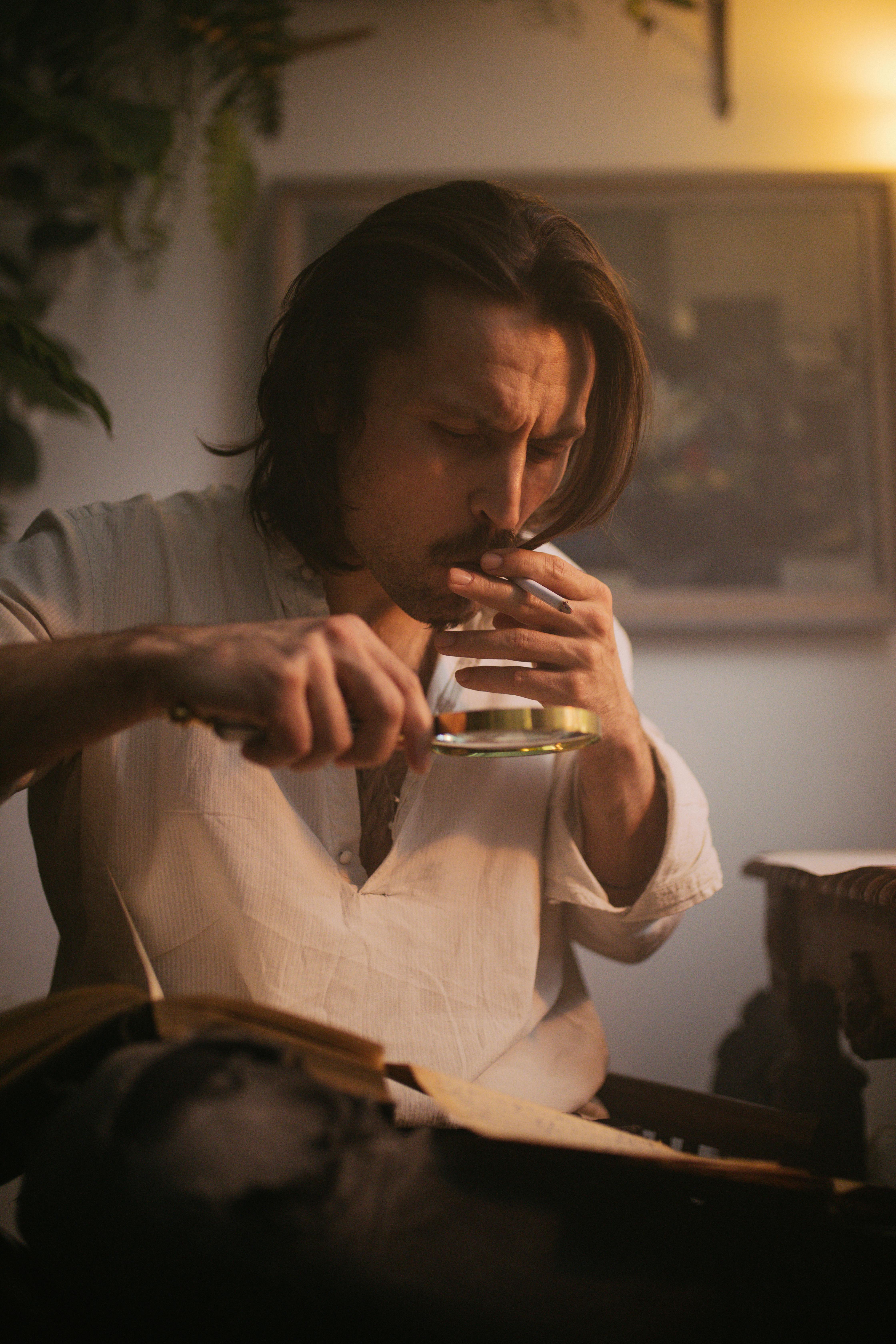 Thoughtful man examining a book with magnifying glass, cigarette in hand, cozy indoor ambiance.