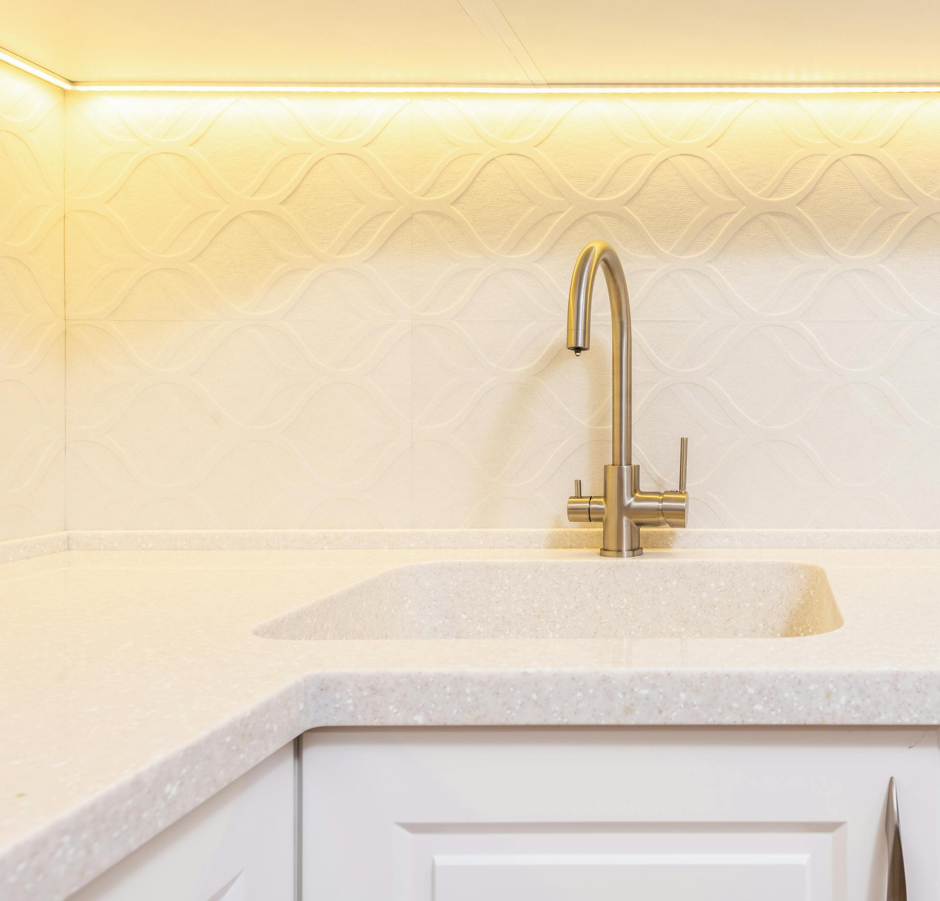 Sink under stainless steel tap against decorative wall illuminated by light in contemporary house kitchen