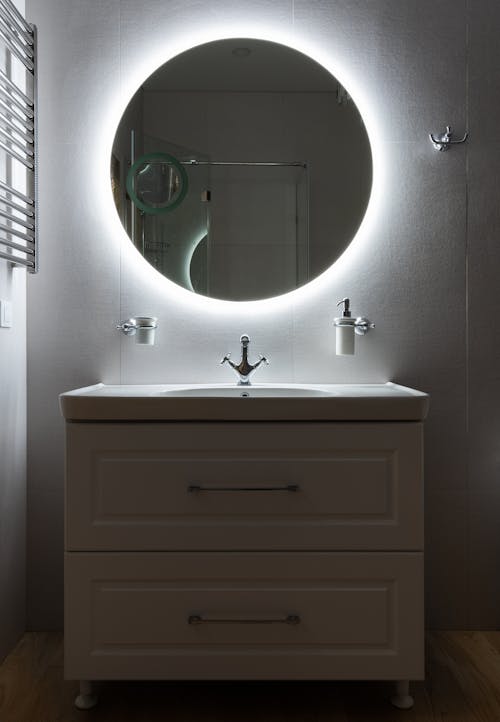 Modern bathroom with washbasin and dispenser bottle under round shaped mirror on light wall in house