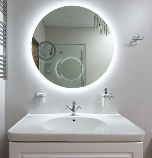Contemporary bathroom interior with washstand under round mirror reflecting shower cabin and chandelier at home