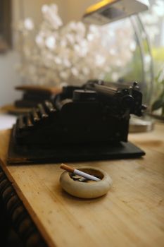 A vintage typewriter and ceramic ashtray with cigarette on a wooden desk, featuring a blurred background.