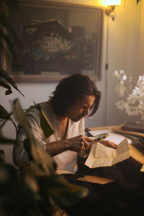 Free Man with magnifying glass examines papers in a dimly lit, artistic study. Warm ambiance. Stock Photo