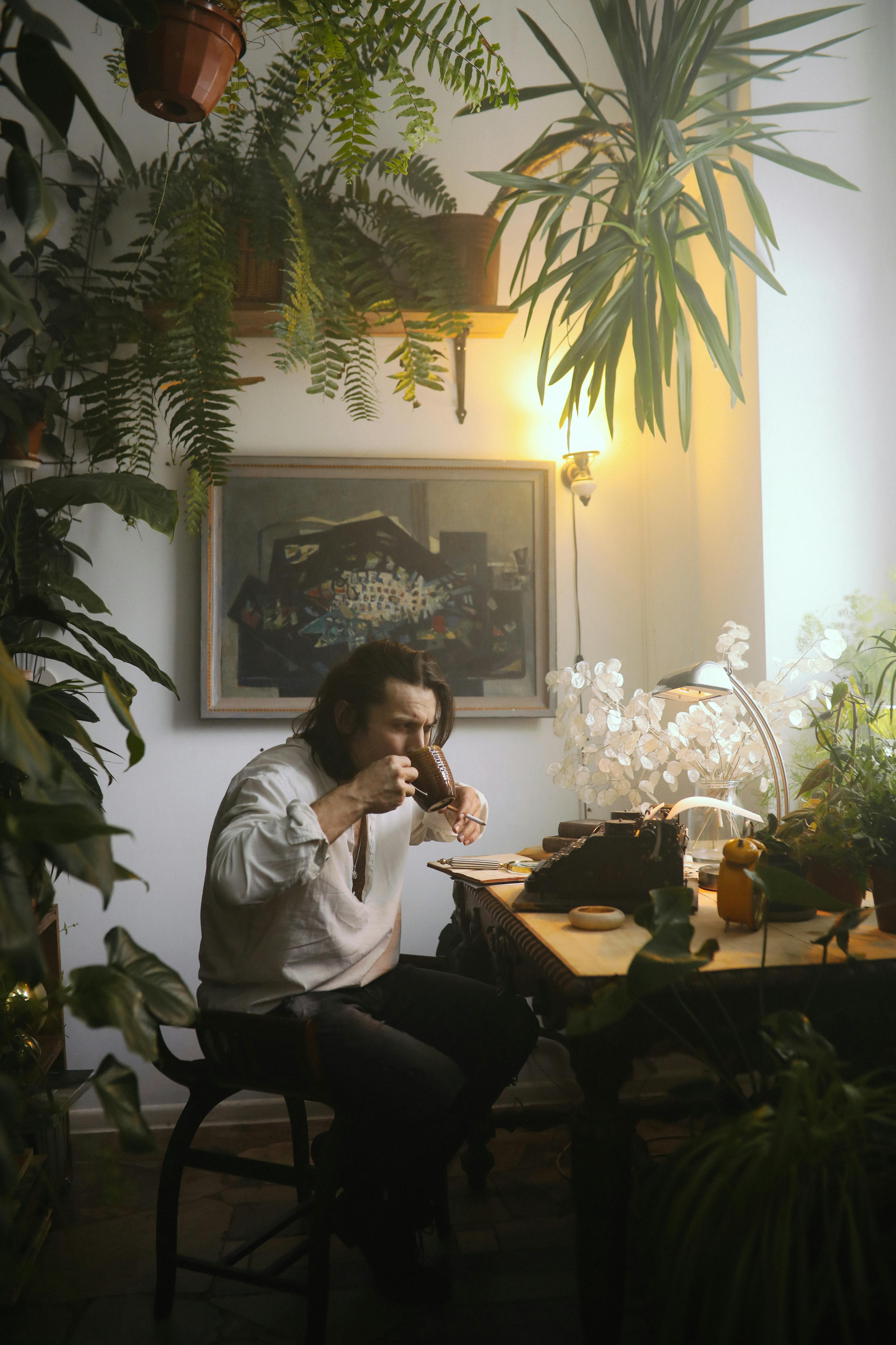 A man enjoys coffee seated at a desk surrounded by lush indoor plants, creating a cozy atmosphere.