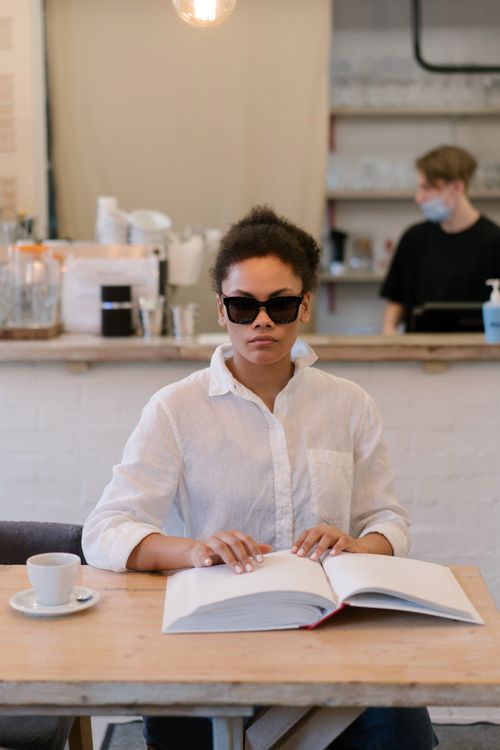 Free Young blind woman wearing sunglasses reading a braille book at a cafe table. Stock Photo