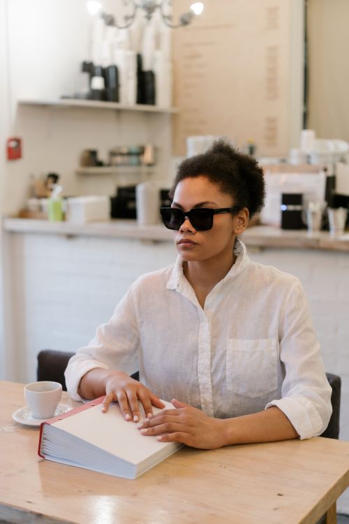 Free African American woman reading Braille book in a cafe, wearing sunglasses, highlighting visual impairment. Stock Photo