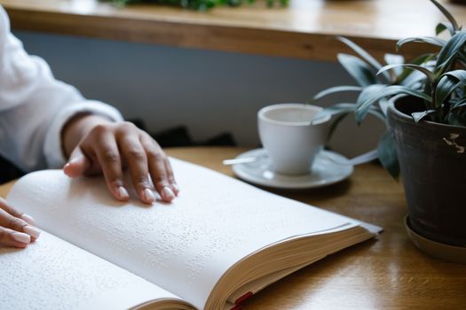 A person reads a Braille book at a wooden table next to a plant and coffee.