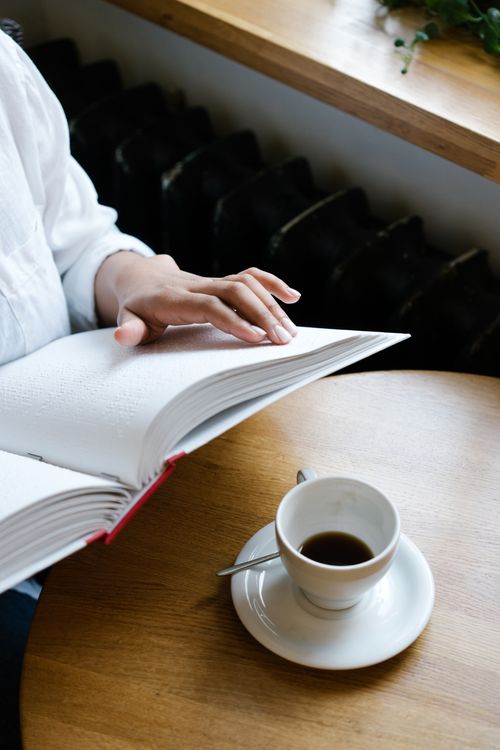 Free A person reading a Braille book beside a cup of coffee on a wooden table. Stock Photo