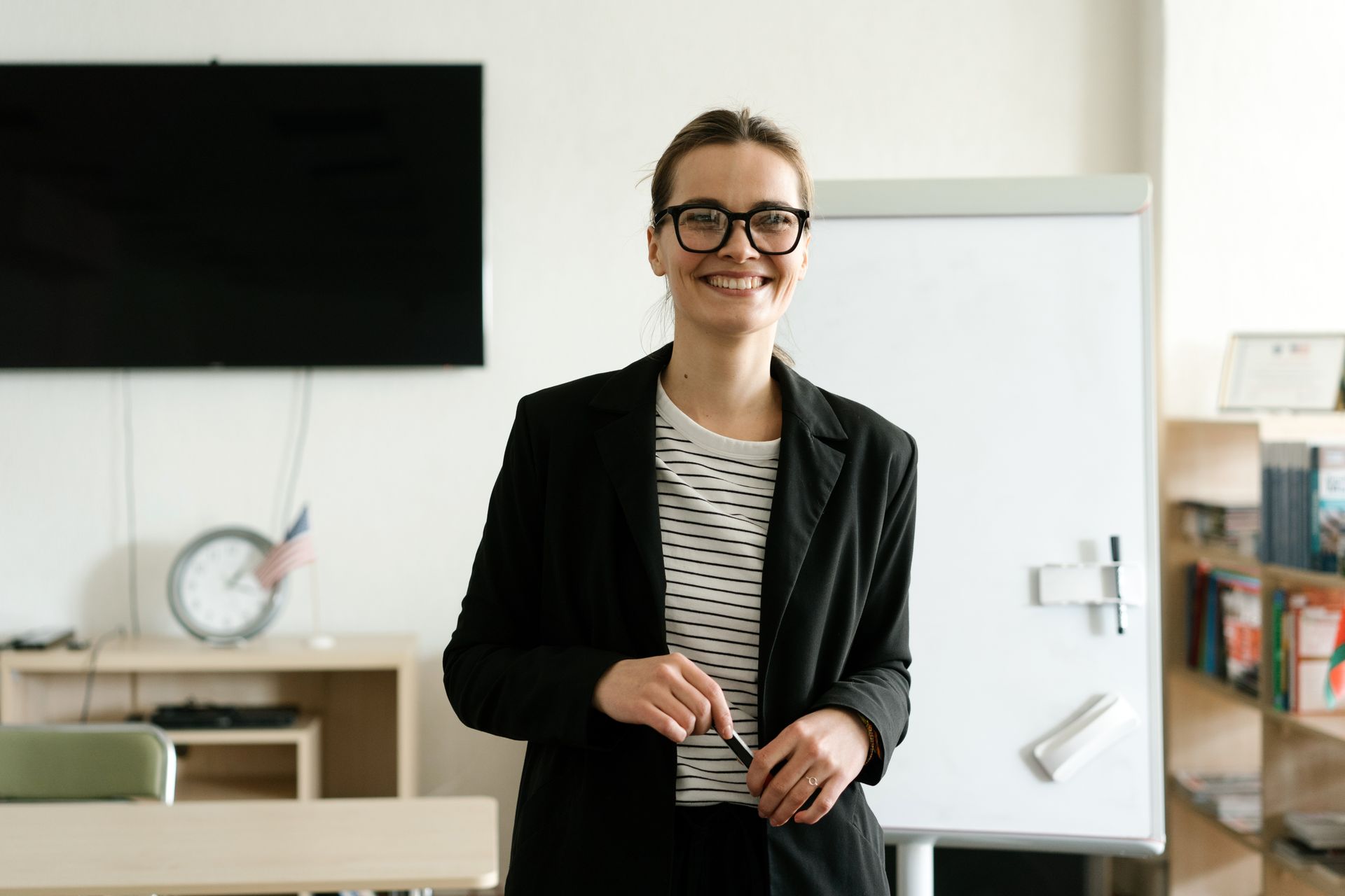 Smiling businesswoman in black blazer with eyeglasses holding a pen in an office setting.