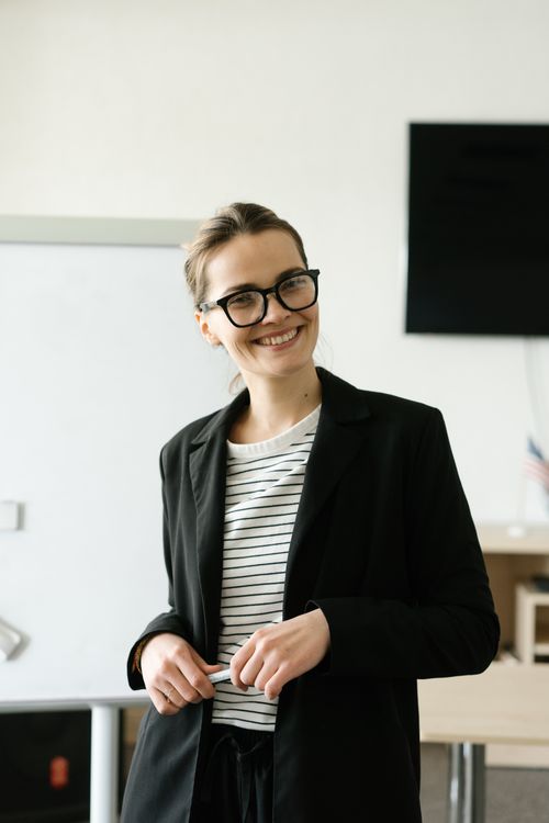 Free Young woman smiling in an indoor setting, wearing glasses and a black blazer. Stock Photo