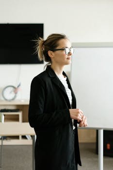 A confident woman wearing a black blazer and glasses in a modern office space.