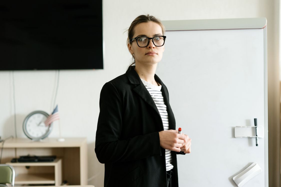 Free Caucasian woman instructor with glasses stands confidently by whiteboard in classroom. Stock Photo