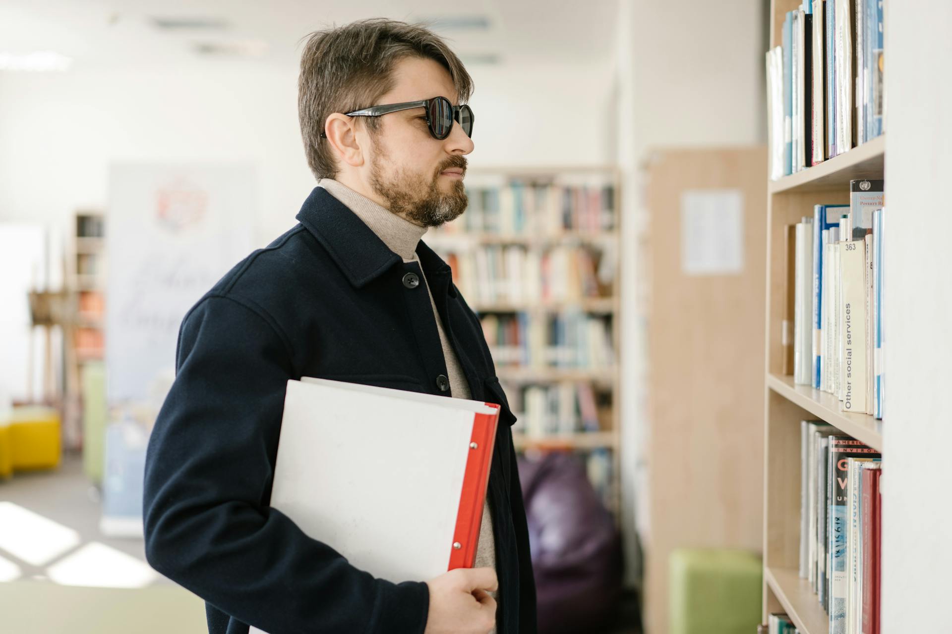 A visually impaired man in sunglasses navigates a library holding a binder, showcasing accessibility and inclusion.