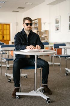 A blind man wearing sunglasses is seated in a classroom, reading a book with braille.