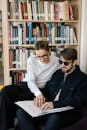 Woman in White Striped Long Sleeve Shirt Sitting Beside a Blind Man Holding and Touching a Braille Book