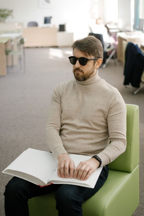 Free A blind man in a library reading a braille book, seated on a green chair wearing sunglasses. Stock Photo