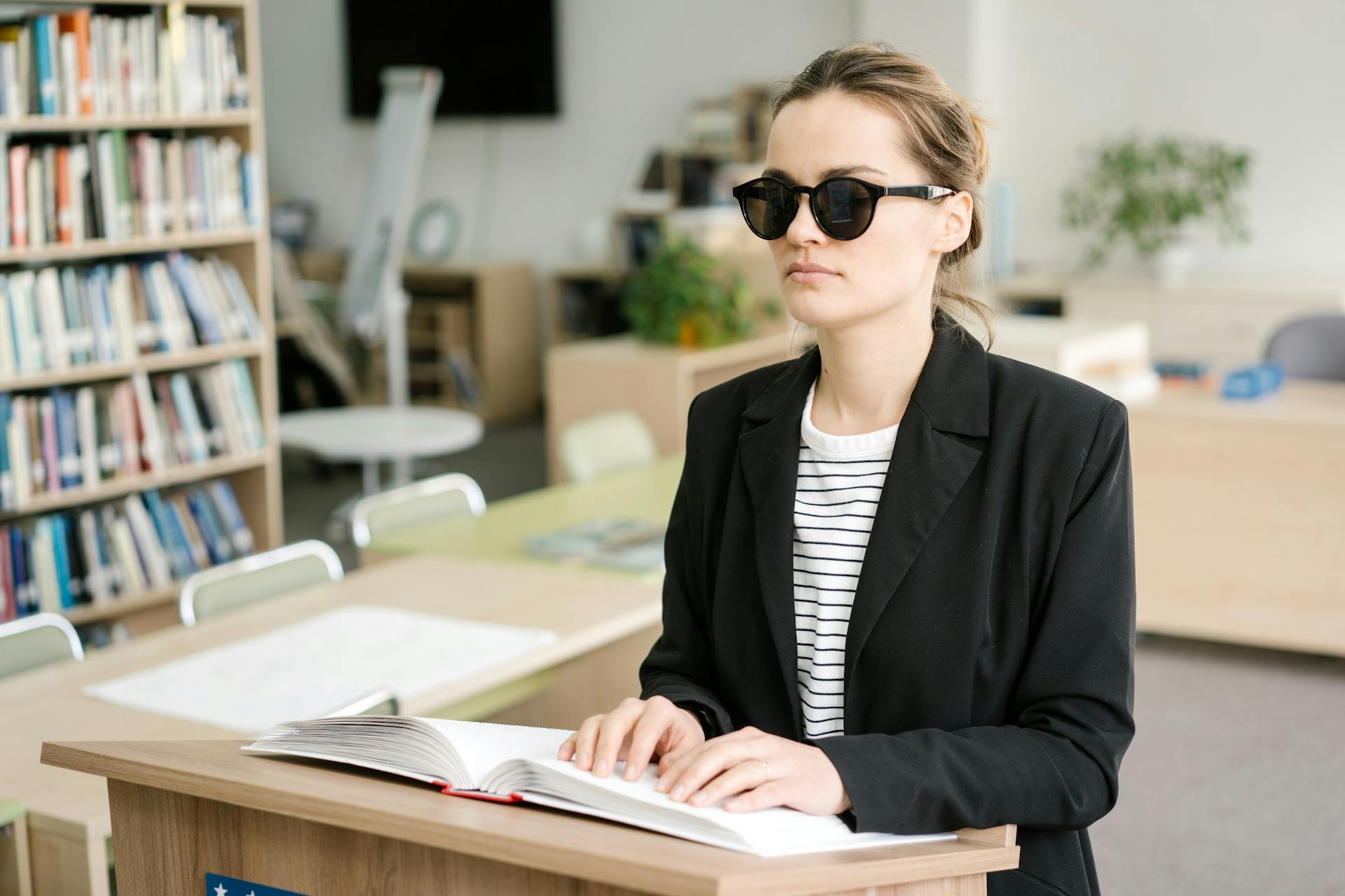 A blind woman in sunglasses reads a Braille book indoors, highlighting disability awareness.