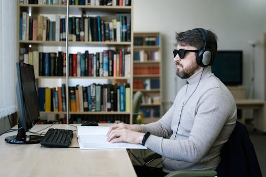 Blind man wearing headphones, working on braille book and computer in a library setting.