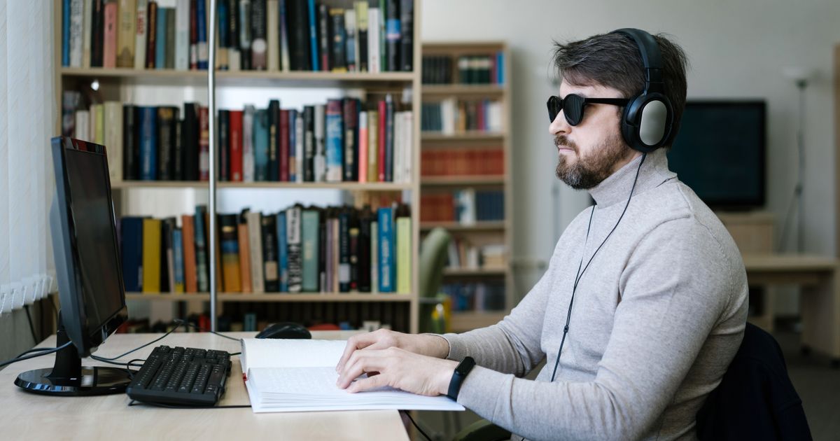 Blinder Mann mit Kopfhörer liest ein Buch in Brailleschrift vor einem Desktop-Computer