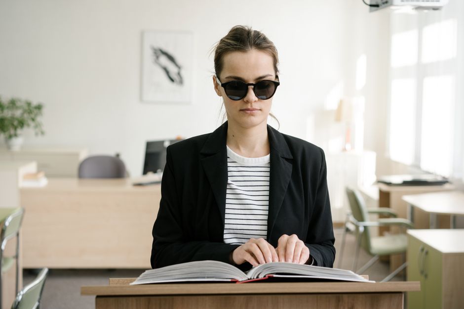 Una mujer ciega con gafas de sol leyendo un libro en braille en un escritorio, simbolizando empoderamiento.