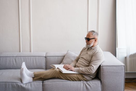 An elderly blind man reading a Braille book while sitting comfortably on a sofa indoors.