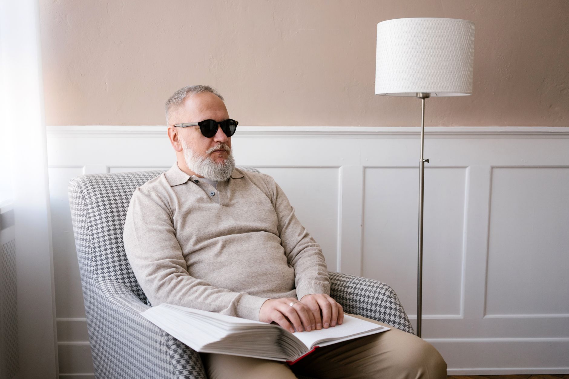 Elderly man with sunglasses reading a braille book indoors, highlighting disability awareness.